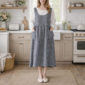 Può includere: Una donna indossa un abito grembiule a quadri blu e bianchi con ampie tasche. Il grembiule è sovrapposto a una camicia bianca a maniche lunghe. L'ambiente della cucina comprende un tappeto, armadi e un fornello.