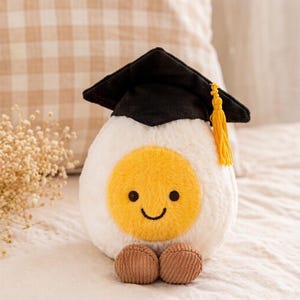May include: A plush toy shaped like a fried egg, featuring a yellow yolk with a smiling face and brown feet. The egg is wearing a black graduation cap with a yellow tassel. The toy is white and soft, perfect for a graduation gift.
