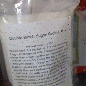 May include: A clear plastic bag of "Double Batch Sugar Cookie Mix" with a colorful polka dot label. The label includes instructions for baking sugar cookies, including ingredients and baking time. The text is in black font.