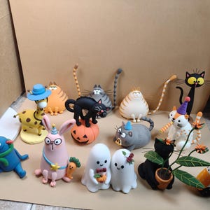 May include: A collection of small, colorful clay figurines featuring various characters including a giraffe, several cats in different poses, a pink rabbit, two ghost figures, and a blue creature, all arranged together on a flat surface.
