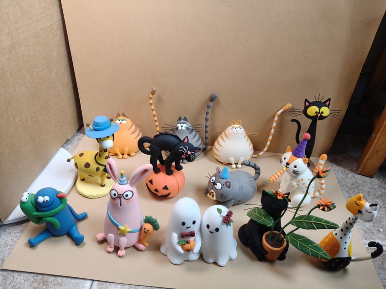 May include: A collection of small, colorful clay figurines featuring various characters including a giraffe, several cats in different poses, a pink rabbit, two ghost figures, and a blue creature, all arranged together on a flat surface.