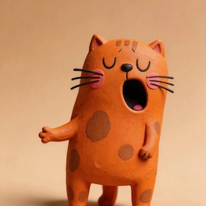 May include: An orange clay figurine of a cat with brown spots, a wide-open mouth, pink circular cheeks, and black whiskers. The cat has closed eyes, small ears, and one arm extended to the side, standing on two short legs.