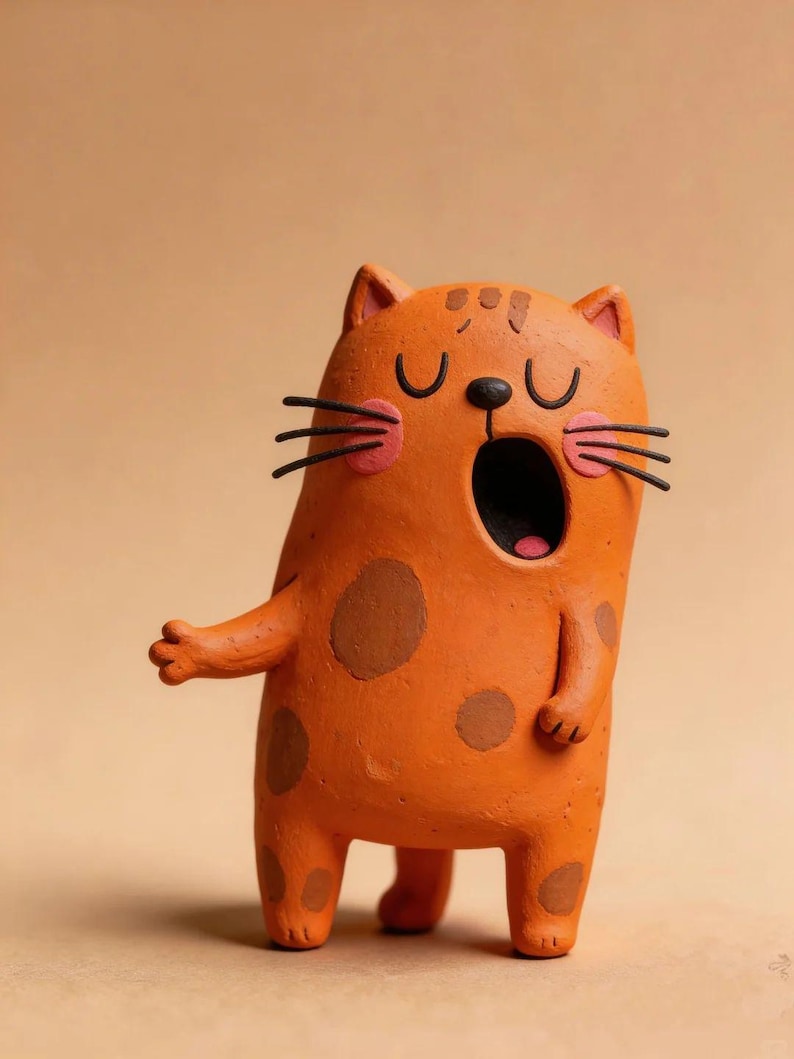 May include: An orange clay figurine of a cat with brown spots, a wide-open mouth, pink circular cheeks, and black whiskers. The cat has closed eyes, small ears, and one arm extended to the side, standing on two short legs.