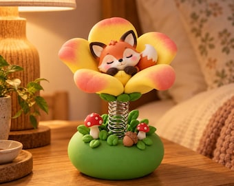 Puede incluir: Una figura decorativa que muestra a un pequeño zorro naranja y blanco durmiendo dentro de una gran flor amarilla y rosa. La flor se asienta sobre una base verde decorada con dos pequeñas setas rojas y blancas y una bellota, conectadas por un resorte metálico.