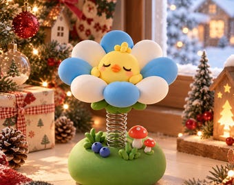 Puede incluir: Un adorno de escritorio montado sobre un resorte que presenta un pollito amarillo con los ojos cerrados en el centro de una flor azul y blanca. La base es verde y está decorada con dos pequeñas setas rojas y blancas, dos bayas azules y hierba.