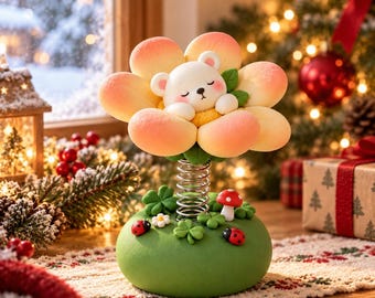 Puede incluir: Una figura decorativa que muestra un oso blanco durmiendo dentro de una flor color melocotón, montada sobre un resorte metálico en una base verde. La base está decorada con hojas de trébol verdes, dos mariquitas rojas, una pequeña flor blanca y una seta roja.