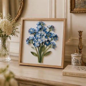 May include: A framed paper quilling artwork depicting a bouquet of blue forget-me-nots. The flowers are made with rolled paper, in shades of blue and white. The artwork is set in a natural wood frame, displayed on a light-coloured surface.