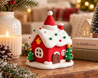 Puede incluir: Una figura de casa de arcilla roja con un techo blanco cubierto de puntos verdes y rojos, una puerta marrón y dos pequeños árboles de Navidad verdes a los lados. El techo presenta un gorro de Papá Noel central y una pequeña chimenea.