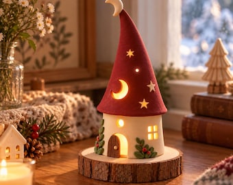 Puede incluir: Una lámpara cónica en forma de casa con techo rojo, rematada con una media luna, estrellas recortadas y una ventana lunar iluminada. La base blanca tiene una puerta arqueada marrón, una pequeña ventana cuadrada y detalles de follaje verde.
