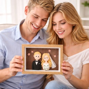 Puede incluir: Un retrato de boda enmarcado con figuras de novios. El novio lleva un esmoquin negro, y la novia un vestido blanco y velo. Acentos de flores rosas rodean las figuras. El marco es de madera clara.