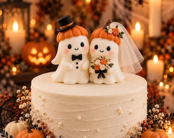 Puede incluir: Un pastel redondo blanco coronado con dos figuras de fantasmas. Un fantasma lleva un sombrero de copa negro y pajarita, mientras que el otro lleva un velo blanco y sostiene un ramo de flores naranjas y blancas. Ambos tienen gorros de calabaza.