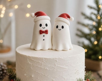 Puede incluir: Dos figuras de fantasmas blancos con ojos negros y bocas sonrientes, cada una con un gorro de Papá Noel rojo con borde blanco. Un fantasma tiene una pajarita roja y dos botones negros. Están colocados sobre un pastel redondo con glaseado blanco.