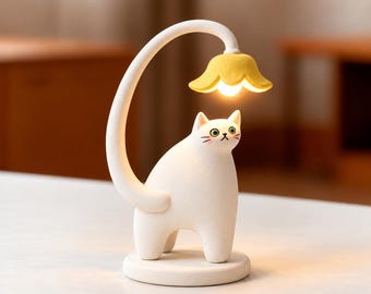 Puede incluir: Una lámpara de mesa con forma de gato blanco y acabado mate liso. La cola del gato se curva hacia arriba para sostener una pantalla amarilla en forma de flor que emite una luz cálida dirigida hacia la cabeza del gato.