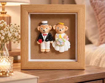 Puede incluir: Un marco cuadrado de madera de roble claro que contiene dos figuras de osos esculpidas vestidas para una boda. El oso de la izquierda lleva un esmoquin negro con pajarita roja y sostiene un corazón rojo. El oso de la derecha lleva un vestido.