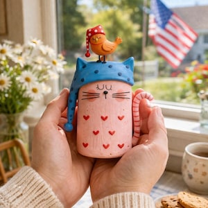 Peut inclure: Une tasse en céramique rose en forme de chat, ornée de cœurs rouges et d'un bonnet de nuit bleu. Un petit oiseau orange avec un chapeau rouge est posé sur la tasse. La tasse a une anse rayée et est tenue à deux mains.