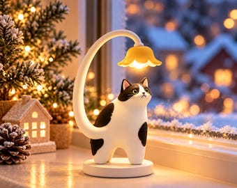 Puede incluir: Una lámpara de mesa con forma de gato blanco y negro con un cuello curvo que sale de su espalda, rematado con una pantalla amarilla en forma de flor que emite un brillo cálido. La lámpara se apoya sobre una base circular blanca lisa.