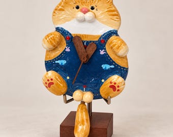 Puede incluir: Un reloj de pared con forma de gato atigrado naranja que lleva un peto vaquero azul con patrones de peces y conejos pequeños. El gato tiene el pecho blanco, la nariz rosa y las almohadillas de las patas rojas. Agujas de madera marrón.