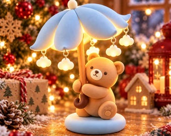 Puede incluir: Una figura de oso de peluche marrón claro que sostiene una lámpara de paraguas azul claro. El paraguas cuenta con dijes blancos iluminados en forma de nube. El oso se asienta sobre una base circular azul claro y emite una luz suave.