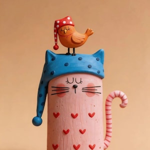 May include: A cylindrical pink cat figurine with red heart patterns, wearing a blue polka-dot nightcap. A small orange bird in a red polka-dot nightcap sits on the cat's head. The cat has a striped tail, closed eyes, and whiskers.