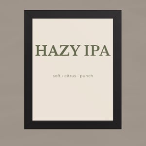 Könnte beinhalten: Ein rechteckiger Wandkunstdruck in einem schwarzen Rahmen mit cremefarbenem Hintergrund. Der Text HAZY IPA ist in großen, dunkelgrünen Großbuchstaben gedruckt, darunter stehen die Wörter soft, citrus und punch in kleineren Kleinbuchstaben.