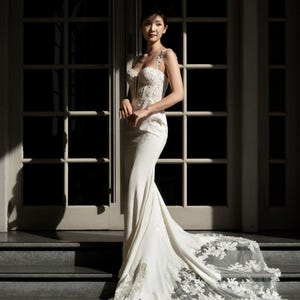 Puede incluir: Un vestido de novia blanco hasta el suelo con un corpiño estructurado con intrincados bordados de encaje floral, tirantes finos y una silueta ajustada que se ensancha en una larga cola de encaje transparente sobre los escalones de piedra.