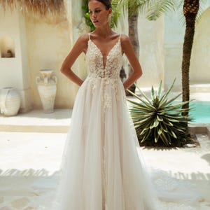 May include: A white wedding dress with a plunging neckline and delicate floral lace detailing. The gown features spaghetti straps and a flowing tulle skirt. The bride is standing outdoors near a pool and palm trees.