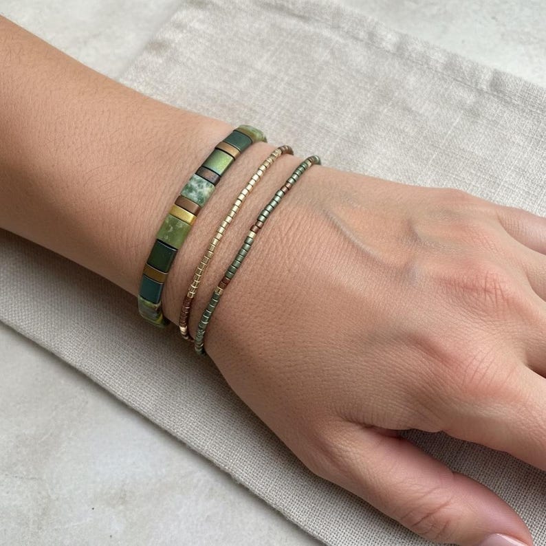 May include: A set of three bracelets worn together. The first is a wide band of square, flat, olive green and metallic bronze beads. The second is a thin strand of small, gold-toned beads. The third is a thin strand of small, dark green beads.