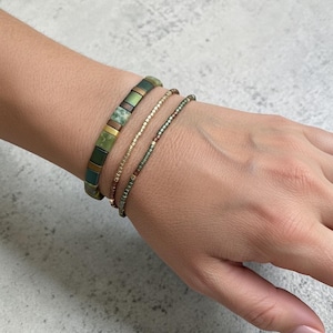 May include: A set of three bracelets featuring a wide band of square green and metallic bronze tiles, alongside two thinner strands composed of small, uniform beads in shades of olive green, metallic gold, and bronze tones arranged in a linear pattern.