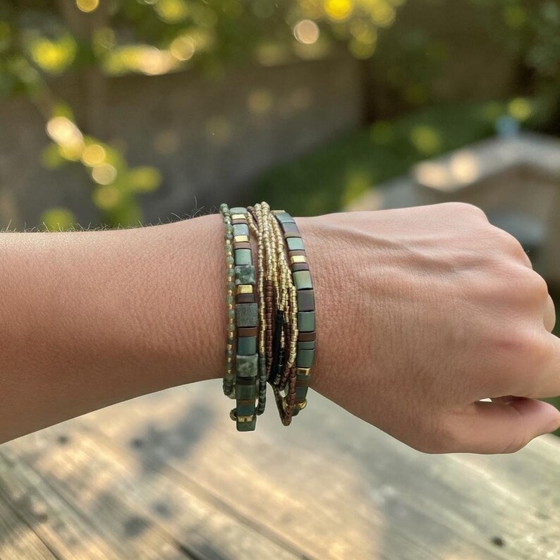 May include: A multi-strand bracelet featuring a combination of small green, brown, and gold-toned beads. The design includes square-shaped matte tiles in shades of sage green and brown, interspersed with thin metallic gold and dark brown seed beads.