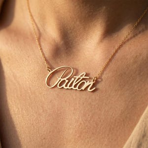 May include: A gold-toned metal necklace featuring a nameplate pendant with the name Paiton written in a cursive script font. The pendant is attached to a delicate, thin gold-toned chain that rests against the skin, showcasing a polished finish.