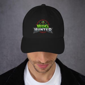 May include: Black baseball cap with the text "BUG HUNTER" in silver and green, featuring a green bug graphic inside a red target. The cap has a curved peak and an adjustable design.