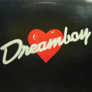 May include: A black square with a red heart and the word "Dreamboy" in white script. The heart is centered, and the script is above and to the right of the heart. The background is black.