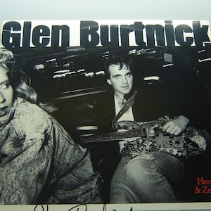 May include: Black and white photo of a man and woman. The man is playing a guitar and the woman is looking at the camera. The album cover is for "Glen Burtnick: Heroes & Zeros".