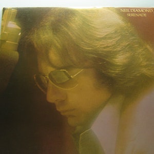 Peut inclure: Pochette d'album pour "Serenade" de Neil Diamond. La couverture présente un profil d'homme portant des lunettes, avec de longs cheveux. La pochette de l'album est présentée sur un support blanc. La pochette de l'album a une teinte sépia.