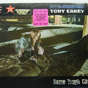 May include: A vinyl record album cover featuring a photo of two people in an urban setting. The album is titled "Some Tough City" by Tony Carey. The album cover also includes a sticker that says "Contains the hits: A Fine Fine Day, The First Day of Summer, A Lonely Life."