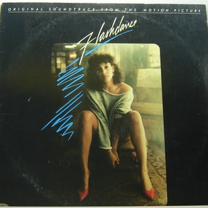 May include: A black vinyl record album cover with the title "Flashdance" in white lettering. The cover features a woman in a grey sweater and red shoes sitting on a couch. The background is a blurry image of a room.