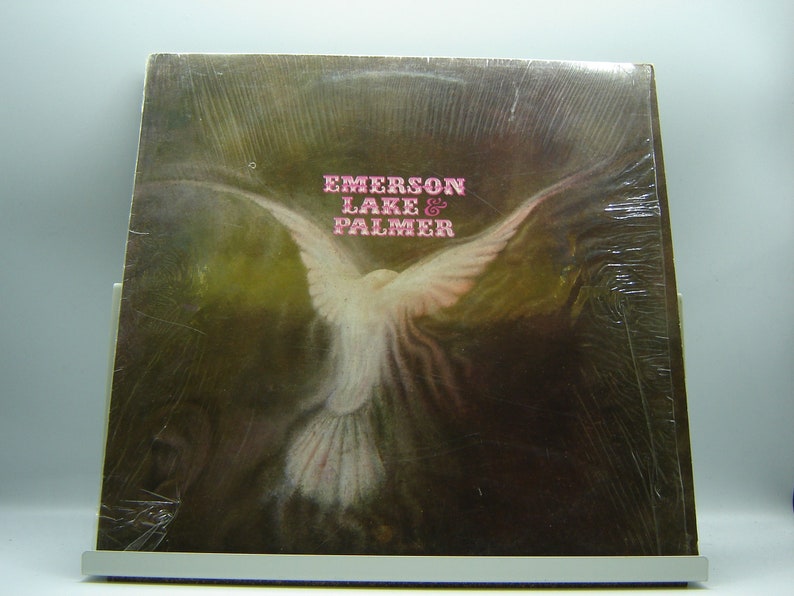 K&ouml;nnte beinhalten: Ein Vintage-Vinyl-Plattencover mit einer wei&szlig;en Taube im Flug vor einem braunen Hintergrund. Der Albumtitel "Emerson Lake & Palmer" ist in rosa Buchstaben gedruckt.