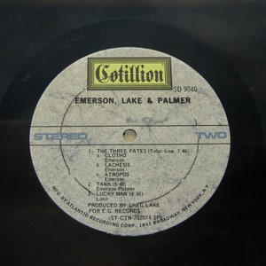 K&ouml;nnte beinhalten: Eine schwarze Schallplatte mit einem wei&szlig;en Label. Das Label zeigt den Text "Cotillion" in einem gelben Kasten, "SD 9040", "Emerson, Lake & Palmer", "Stereo", "Zwei" sowie die Songtitel und K&uuml;nstler. Das Label zeigt auch den Text "MFG. BY ATLANTIC RECORDING CORP., 1841 BROADWAY, NEW YORK, N.Y.", "PRODUCED BY GREG LAKE FOR E.G. RECORDS (ST-CTN-702074 SP"
