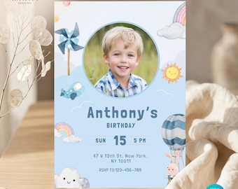 Puede incluir: Una invitación de cumpleaños azul claro con una foto de un niño sonriente. La invitación incluye el nombre "Anthony's" y los detalles de la fiesta: fecha, hora y lugar. Los elementos decorativos incluyen un arcoíris, nubes y un globo aerostático.