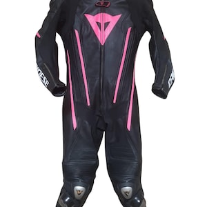 Puede incluir: Traje de carreras de moto negro con detalles en rosa. El traje tiene una cremallera completa, protecciones en rodillas y hombros, y la marca "DAINESE" impresa en las mangas. Un logotipo rosa est&aacute; en el pecho.