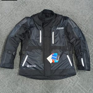 Puede incluir: Chaqueta de moto negra con detalles blancos, múltiples bolsillos y el logo "KLIM". La chaqueta presenta una etiqueta "GORE-TEX" y correas ajustables. La chaqueta está tendida sobre una superficie gris.