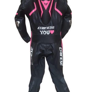 Puede incluir: Traje de carreras de moto de cuero negro con detalles en rosa y el texto "DAINESE YOU". El traje presenta un dise&ntilde;o aerodin&aacute;mico con acolchado protector en hombros y codos. Dise&ntilde;ado para la seguridad y el rendimiento.