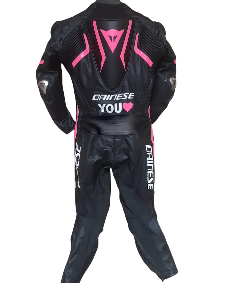 Puede incluir: Traje de carreras de moto de cuero negro con detalles en rosa y el texto "DAINESE YOU". El traje presenta un dise&ntilde;o aerodin&aacute;mico con acolchado protector en hombros y codos. Dise&ntilde;ado para la seguridad y el rendimiento.