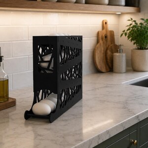 May include: A black, modern egg holder with a decorative, openwork design. The holder is on a marble countertop in a kitchen setting. Several white eggs are visible inside the holder. Other kitchen items include plants, cutting boards, and a soap dispenser.