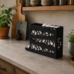 May include: A black metal egg holder with a decorative geometric design, filled with white eggs. The holder sits on a kitchen countertop, alongside a potted plant, a bottle of oil, and other kitchen items.