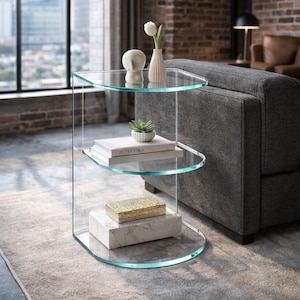 May include: A clear glass side table with three curved shelves. The top shelf holds a white vase with a single white flower and a small, white, abstract sculpture. The middle shelf displays a stack of books and a small potted plant. The bottom shelf has a marble base with a gold box.
