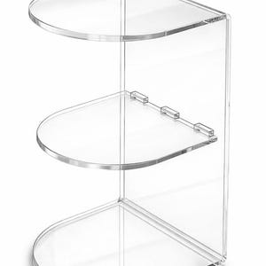 May include: Clear acrylic corner shelf with three rounded shelves. The shelves are connected by a clear vertical support. The design is modern and minimalist, suitable for displaying items in a bathroom or living space.