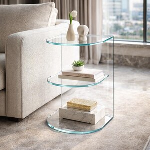 May include: A clear glass side table with three curved shelves. The top shelf holds a small vase with a white flower and a white sculpture. The middle shelf has books and a small plant. The bottom shelf has a marble box with a gold box on top.