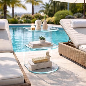 May include: A clear glass side table with three curved shelves. The top shelf holds a candle, a white head sculpture, and a small vase with a white flower. The middle shelf has a small succulent and two books. The bottom shelf has a decorative box and a bust.