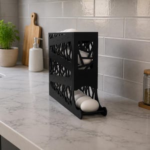 May include: A black, vertical egg holder with a decorative, openwork design. The holder is on a marble countertop and contains several white eggs. Kitchen decor.